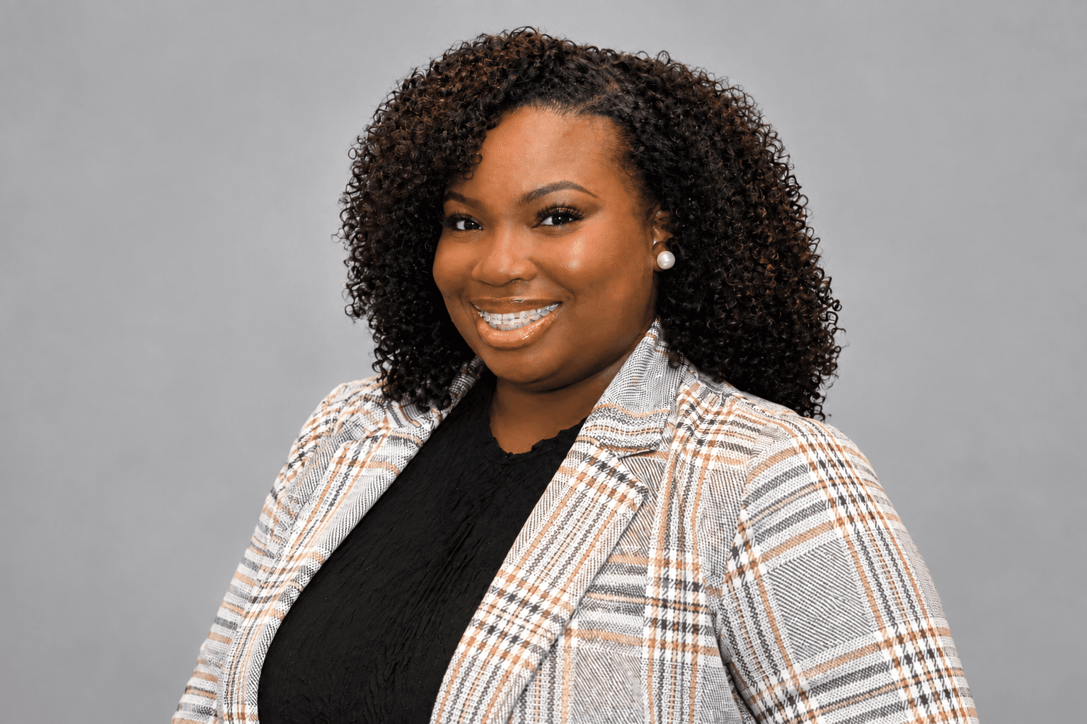 Smiling Black woman with curly hair and braces wearing a plaid blazer against gray background.