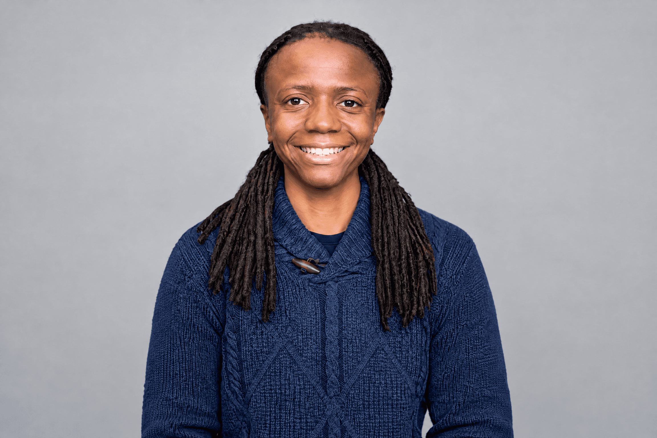 Smiling person with dark dreadlocks wearing a blue cable-knit sweater against a gray background.